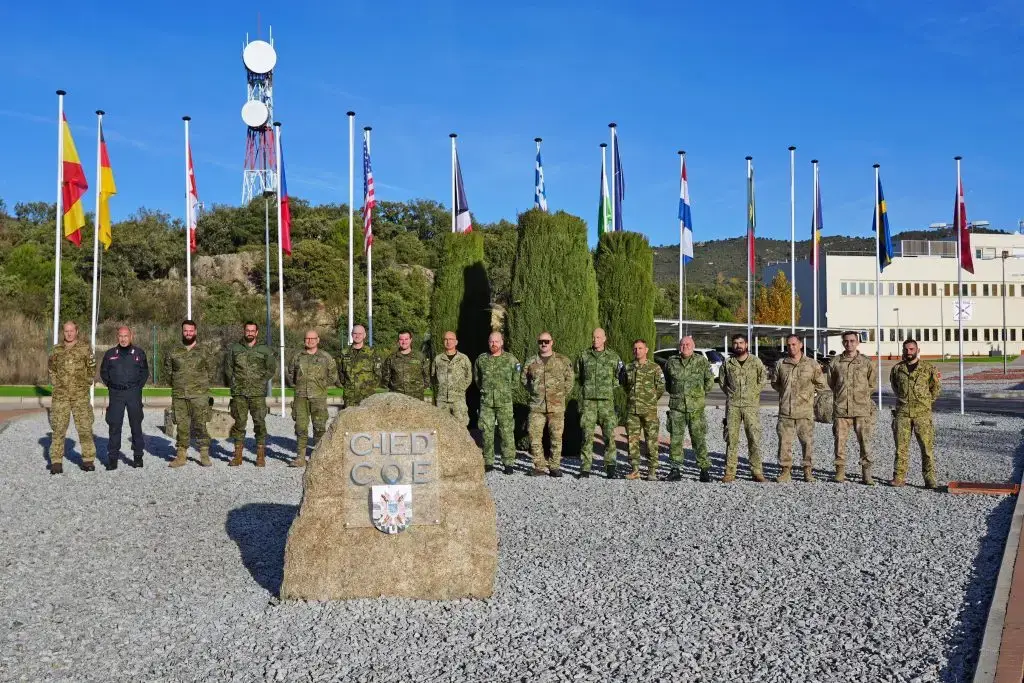 Group of uniformed military personnel standing in formation behind the C-IED COE emblem, with national flags displayed at the headquarters.